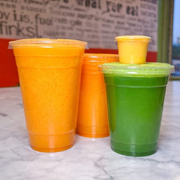 Four plastic cups of vibrant orange and green smoothies and juices on a marble counter.