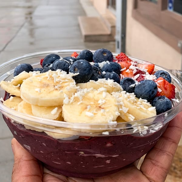 Hand holding an acai bowl topped with sliced bananas, blueberries, strawberries, and shredded coconut.