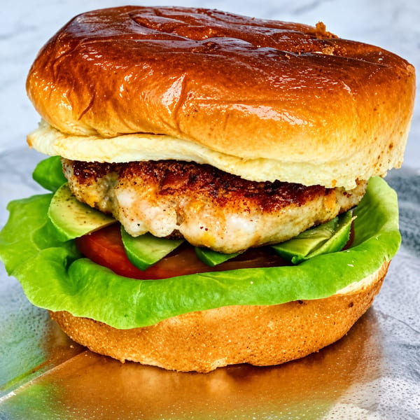 Grilled chicken sandwich on a brioche bun with lettuce, tomato, avocado, and a fried egg.